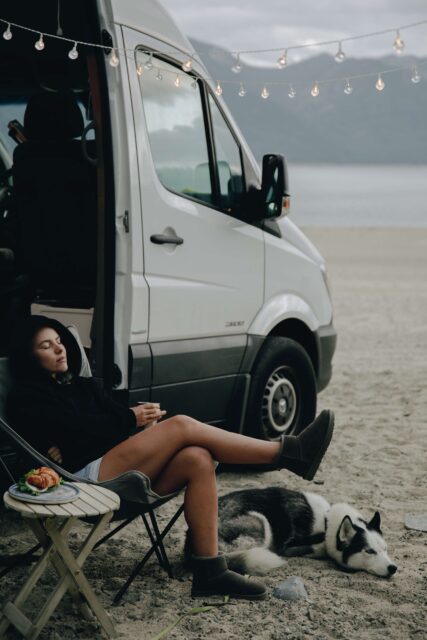 A motorhome on the beach. A woman in a sweater is sitting in a camping chair in front of it. A dog lies at her feet.